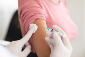 Mulher recebendo aplicação de vacina no braço. O profissional que aplica a vacina está segurando a seringa e utiliza luvas